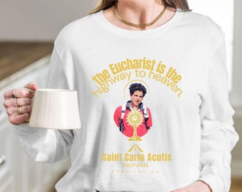 Puede incluir: Camiseta blanca de manga larga con un gráfico de San Carlo Acutis sosteniendo una custodia, con el texto "The Eucharist is the highway to heaven." y "Saint Carlo Acutis 09.07.2025 Pray for us."