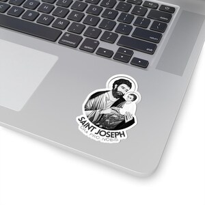 May include: A black and white sticker featuring an illustration of Saint Joseph holding the baby Jesus. The sticker has the text "SAINT JOSEPH ORA PRO NOBIS" in a stylized font. The sticker is placed on a laptop.