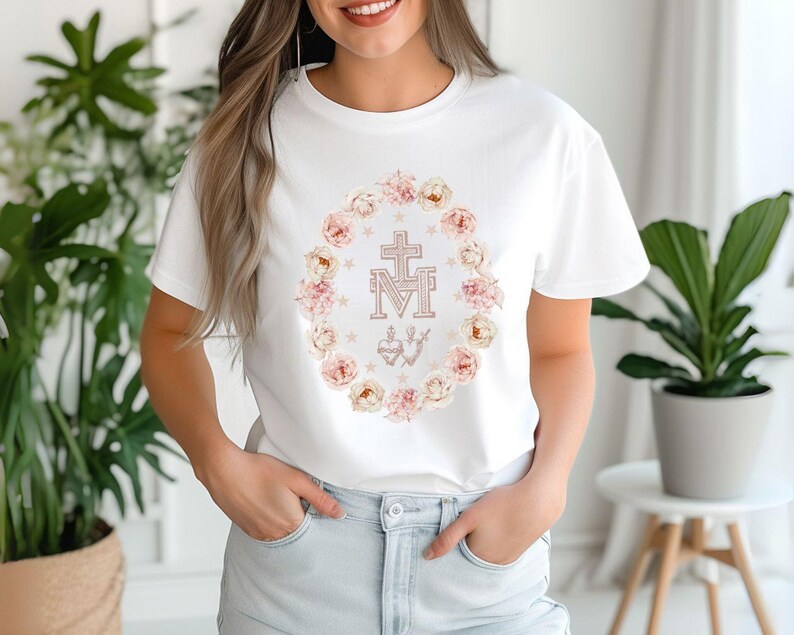 May include: White t-shirt featuring a floral wreath design with a cross and the letters "M" and "T" in the center. The wreath includes pink and cream-colored roses and small stars. The shirt is a classic crew neck style.