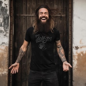 Puede incluir: Un hombre con el pelo largo y barba lleva una camiseta negra con el texto "Your design" y un pequeño gráfico. Está sonriendo y mirando a la cámara.