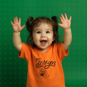 Puede incluir: Un niño pequeño con una camiseta naranja con el texto "Your design here" y un pequeño gráfico de un animal. El niño sonríe y tiene los brazos levantados en el aire.