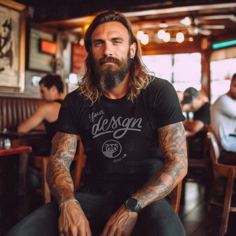 May include: A man with a long beard and tattoos is wearing a black t-shirt with the text "Your design here" and a small graphic of a raccoon.