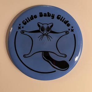 May include: A blue disc with a black outline and a black illustration of a sugar glider with the text "Glide Baby Glide" in a black retro font.
