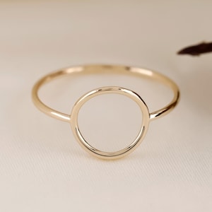 May include: A delicate gold ring featuring a circular design. The ring has a thin band and a hollow circle as its centerpiece. The ring is set against a soft, neutral background, highlighting its simple elegance. A minimalist piece of jewelry.