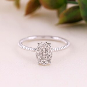 May include: A silver-toned ring featuring a textured, beaded band and an oval-shaped centerpiece cluster of small, clear, sparkling stones. The setting is metallic and polished, showcasing a delicate design with a central oval arrangement of gems.