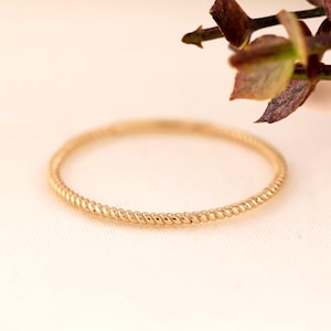 May include: A thin, circular gold ring featuring a continuous twisted rope texture along the entire band. The metal has a polished, reflective finish, creating a delicate and uniform appearance for this simple piece of jewelry.