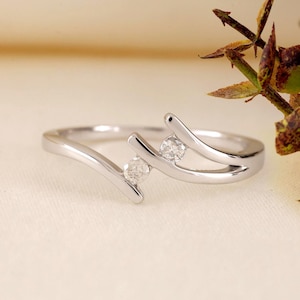 May include: A silver ring with two clear, round gemstones. The ring has a unique design with curved bands that cradle the stones. The ring is set against a soft, off-white background.