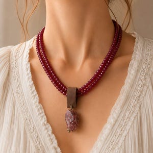 May include: A multi-strand necklace featuring deep red, faceted beads arranged in two rows. A dark brown leather loop connects the strands to a single, irregular, rough-cut purple stone pendant that hangs centrally from a small metal ring.