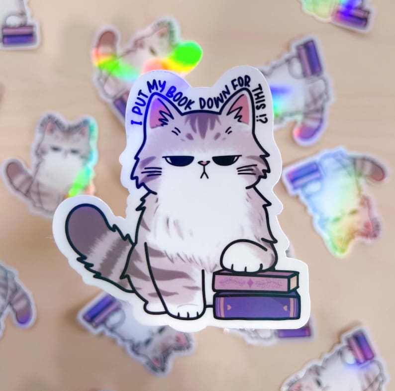 May include: A white and gray cat with a grumpy expression sits on a stack of purple books. The cat is holding a book with its paw and the text "I put my book down for this?" is written above the cat.