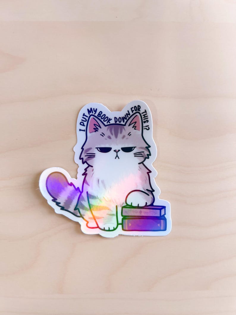 May include: A holographic sticker of a cartoon cat with a grumpy expression. The cat is sitting on top of two books and has a speech bubble above its head that says "I put my book down for this?"