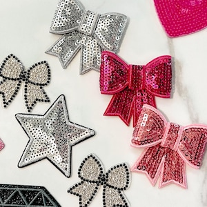 Puede incluir: Surtido de parches de lentejuelas en varias formas, incluyendo lazos, una estrella y un diamante. Los parches presentan lentejuelas plateadas, rosas y negras, algunos con contornos negros. Estos parches decorativos son ideales para embellecer ropa o accesorios.