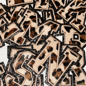 May include: A collection of leopard print fabric letters with black glitter trim. The letters are in various sizes and orientations, with a tan and brown spotted pattern. The letters are likely for crafting or personalization projects.