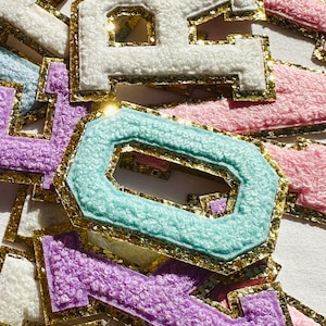 Puede incluir: Primer plano de varias letras de fieltro con ribete de purpurina dorada. Las letras son de varios colores, incluyendo rosa, morado, azul y blanco.