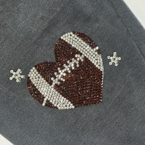 May include: Close-up of a gray fabric with a heart-shaped design. The heart is embellished with brown and silver rhinestones, resembling a football. Two small, silver rhinestone starbursts are also present.