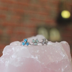 Könnte beinhalten: Drei silberne Ringe mit Türkis- und Opal-Steinen, die in Blumenform gefasst sind, auf einem rosa Rosenquarz-Stein ausgestellt.