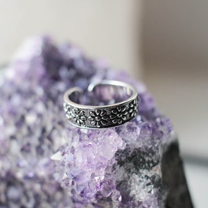 Könnte beinhalten: Ein silberner Ring mit floralem Design, der auf einem Bett aus violetten Amethystkristallen liegt.