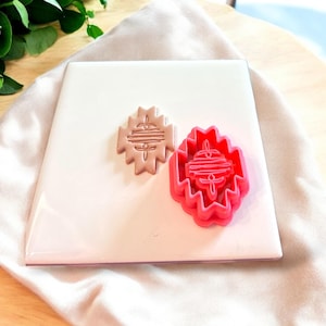 May include: Two cookie cutters, one pink and one brown, with a geometric design. The cutters are on a white surface.