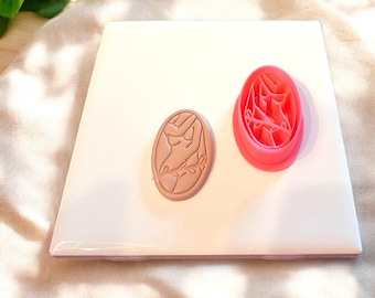 May include: Two pink and brown oval-shaped cookie cutters. The cutter on the left is brown and features a stylized female figure. The cutter on the right is pink and features a stylized floral design.