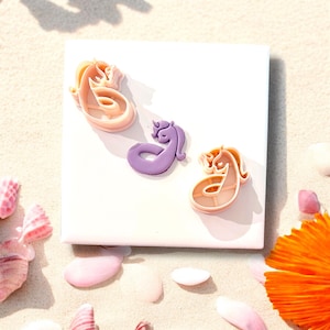 May include: Three whimsical seahorse-shaped cookie cutters in shades of pink and purple rest on a white square. The cutters are arranged on a sandy surface with seashells and flower petals scattered around. The seahorse cutters are a fun addition to any baking collection.
