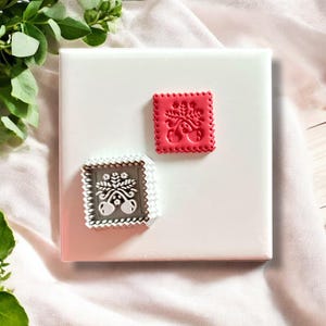 Puede incluir: Dos cortadores de galletas de plástico blanco con un diseño en relieve de una rama de cerezo con hojas y cerezas. El cortador de la izquierda está sobre una superficie blanca. El cortador de la derecha es rojo.