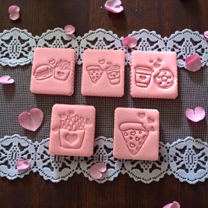 May include: Pink square cookie stamps with food-themed designs, including a burger, fries, pizza, coffee, and a donut. The stamps are arranged on a white lace runner with pink heart-shaped petals scattered around.