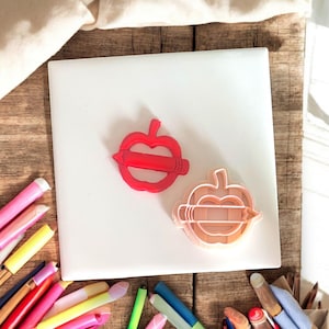 May include: Two apple-shaped cookie cutters, one red and one peach-coloured, sit on a white surface. The cutters have a pencil design through the centre. A collection of colourful art supplies surrounds the white surface.