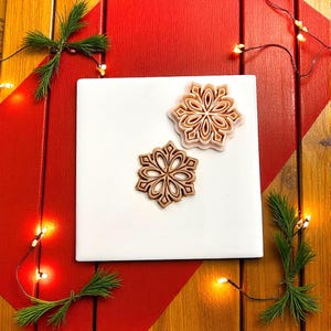 Può includere: Due formine per biscotti a forma di fiocco di neve in marrone chiaro e rosa, appoggiate su un quadrato bianco. Le formine sono circondate da rametti di pino e una stringa di lucine, su uno sfondo rosso e in legno. Festivo e stagionale.