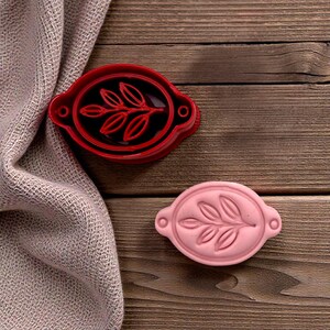 May include: Two oval-shaped cookie cutters with a leaf design. One is red, the other is pink. The cutters are on a wooden surface next to a folded beige cloth. The leaf design is in the center of the oval.