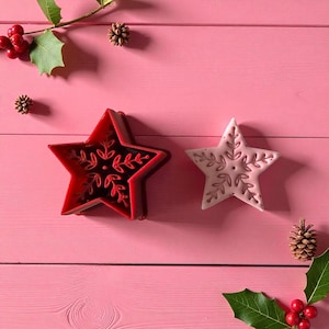 May include: Two star-shaped Christmas decorations. One is red with a snowflake design, and the other is light pink with a similar pattern. The stars are on a pink wooden surface, with holly and pinecones scattered around.