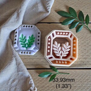 May include: Two octagonal soap molds. One is light gray with a green leaf design, the other is peach-colored with a leaf design. The molds are approximately 1.33 inches (33.93mm) in size. The molds are on a wooden surface.