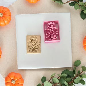 Puede incluir: Un cortador de galletas de plástico rosa y una galleta estampada con un diseño de murciélago. El cortador de galletas es rectangular con un diseño de murciélago y luna. La galleta estampada es de color marrón claro con el mismo diseño de murciélago.