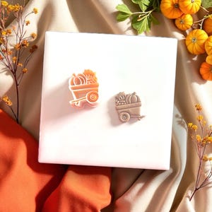 May include: Two pumpkin-themed cookie cutters. One is light orange, the other is tan. Both cutters depict a wagon filled with pumpkins and gourds. The cutters are on a white surface, with orange and yellow fall foliage in the background.