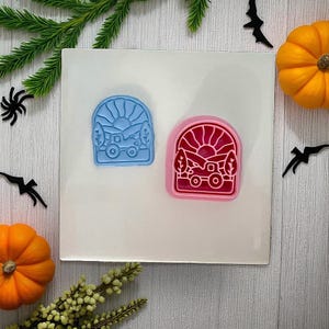 May include: Two colorful silicone molds with a nature-themed design. One is blue, the other is pink. Each mold features a sun, tractor, trees, and mountains. The molds are on a white surface, surrounded by Halloween decorations and greenery.
