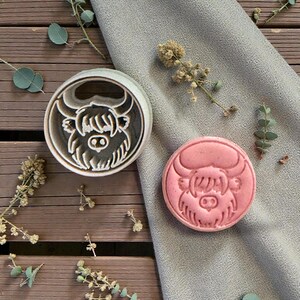 May include: A round cookie cutter and a pink biscuit, both featuring a Highland cow design. The cutter is light grey with a detailed cow face, while the biscuit is pink with the same embossed design. The items are on a wooden surface.
