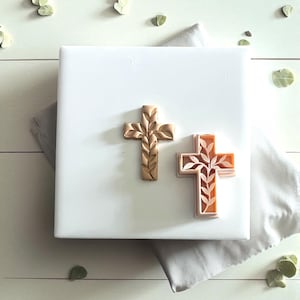 May include: Two decorative cross-shaped molds. One is a solid tan color with leaf-like details. The other is a light orange color with a leaf design. Both are on a white surface with a gray cloth and scattered green leaves.