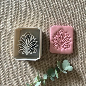 May include: A white plastic cookie cutter with a floral design next to a pink clay impression of the same design. The floral design has a central petal shape with curved leaves and swirls. The items are on a textured beige surface.