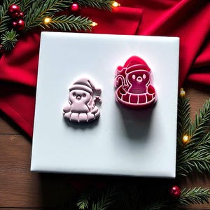 May include: Two pink Christmas-themed cookie cutters shaped like a Santa Claus penguin. One is a lighter pink, and the other is a darker pink. The cookie cutters are on a white surface, with red fabric and Christmas greenery in the background.