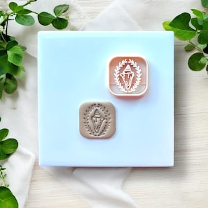 May include: A light blue square with a white cloth and green leaves in the background. Two square wax seal stamps are displayed. One is a light pink color with a white design of a shell and laurel wreath. The other is a tan color with the same shell and wreath design.