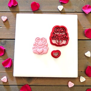 May include: A pink frog-shaped cookie cutter and a red frog-shaped cookie cutter with a heart. A small red heart-shaped cookie cutter is also present. The items are on a white surface, surrounded by pink rose petals and heart-shaped candies.
