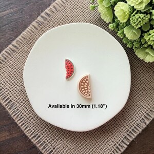 May include: Two fruit-themed charms, one a red and green watermelon slice, the other a tan and brown orange slice, rest on a white plate. The text "Available in 30mm (1.18")" is below the charms. The plate sits on a burlap surface.