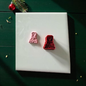 May include: Two cookie cutters in the shape of a nutcracker's face. One is pink, the other is red. The cutters are on a white surface, with a green background and Christmas decorations.