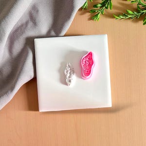 May include: Two small, decorative items sit on a white surface. One is a light gray, abstract shape, and the other is a pink, irregular shape with a design inside. The background is a light brown surface with a gray cloth and green leaves.