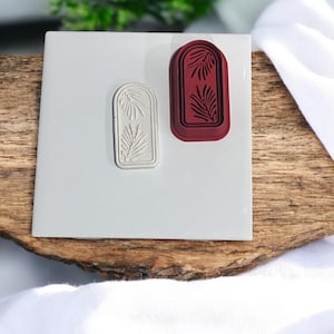 Puede incluir: Dos sellos de goma rectangulares con un diseño de hojas. Un sello es rojo y el otro es blanco.