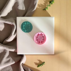 May include: Two round, decorative stamps with geometric designs. One stamp is teal, the other is pink. The stamps are on a white square surface, with a beige background and a draped fabric in the upper left corner. Green sprigs are placed around the stamps.