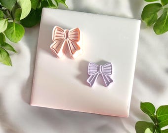 May include: Two bow-shaped cookie cutters. One is light pink, the other is lavender. The cutters are on a white and pink surface, surrounded by green leaves. These are ideal for baking and crafting.