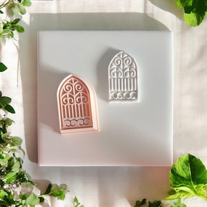 May include: Two decorative window-shaped cookie cutters. One is a light peach color, and the other is white. Both have intricate scrollwork and vertical bars. The cutters are on a white surface, with green foliage in the corners.