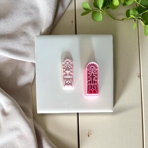 May include: Two pink cookie cutters in the shape of stained glass windows. One is a light pink, the other is a darker pink. The cutters are on a white surface, with a gray cloth and greenery in the background.