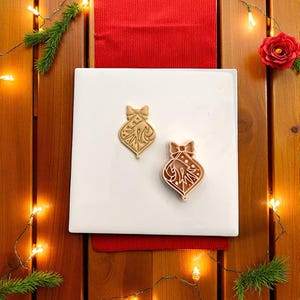 以下が含まれることがあります： クリスマスオーナメント型のクッキーカッターが2つ。1つはタン色、もう1つはピンク色。どちらも上部にリボンがあり、装飾的な切り抜きデザインが施されています。白い表面に赤いランナーがあり、クリスマスの装飾とライトに囲まれています。