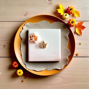 May include: A fall-themed image featuring a white square tile with two leaf-shaped cookie cutters. The cutters are in shades of orange and brown. The tile sits on a tan plate with miniature pumpkins and fall leaves.