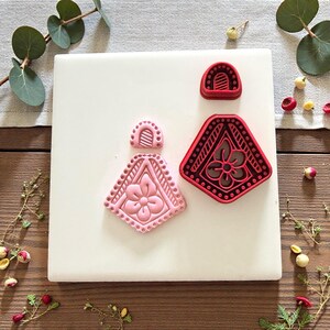 May include: Three decorative cookie cutters in the shape of earrings. One is pink, and two are red. The pink cutter has a floral design. The red cutters have a geometric design. The cutters are on a white surface.